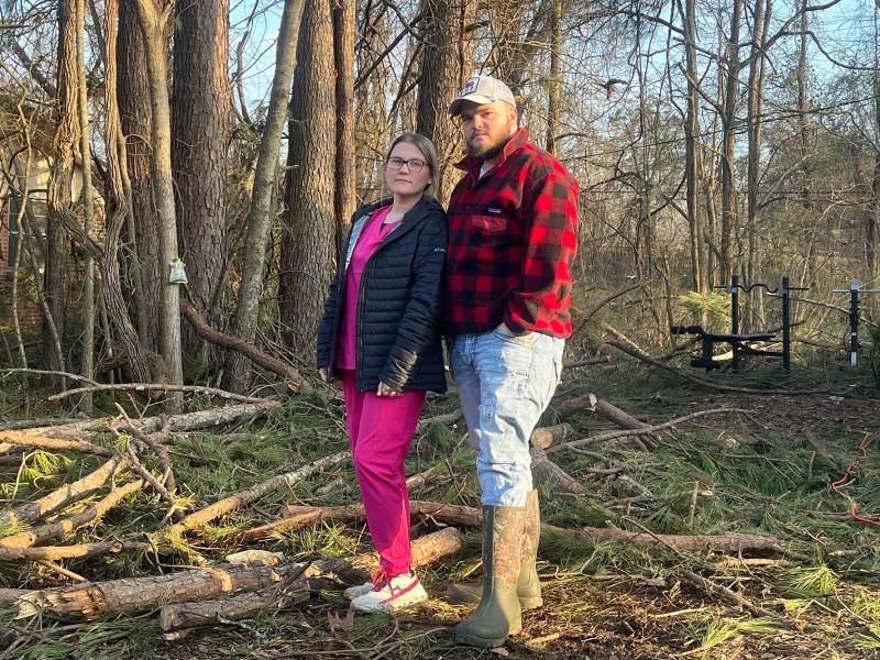 A couple wear jackets and stand in a yard filled with fallen tree debris