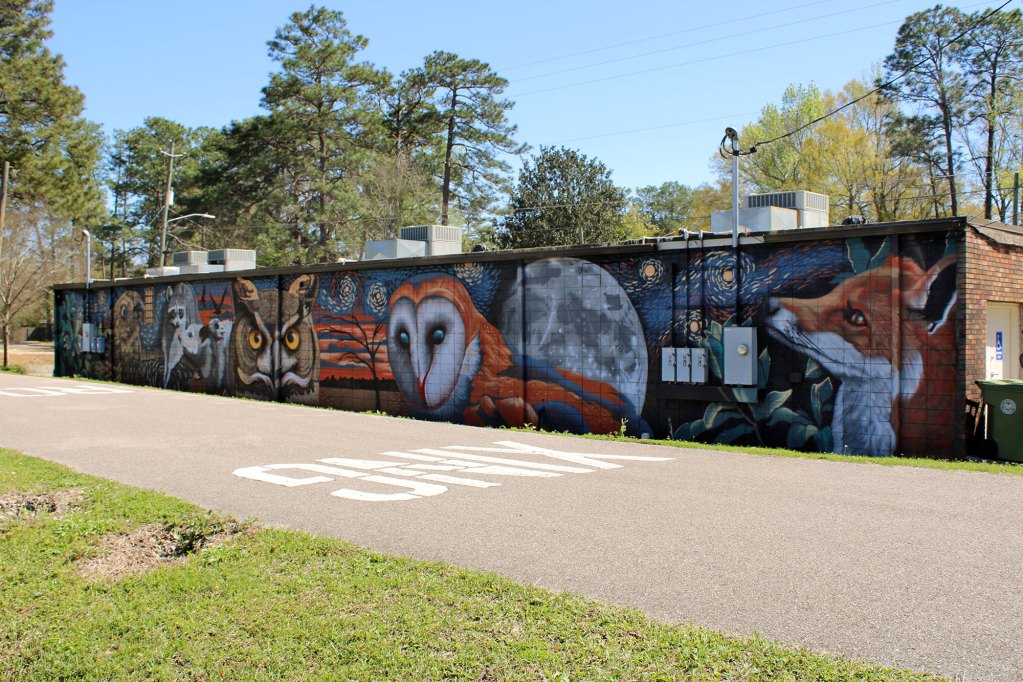 A mural spans the full back of a brick building featuring nocturnal creatures like owls and foxes