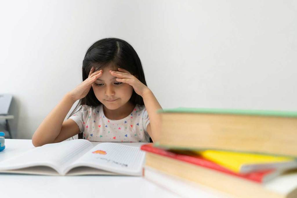 A young girl seated at a table reading. Her head is resting on her hands.