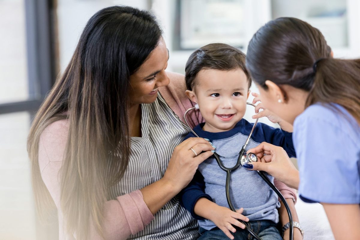 A woman with a smiling child while another woman holds up a stethoscope to the child’s chest.