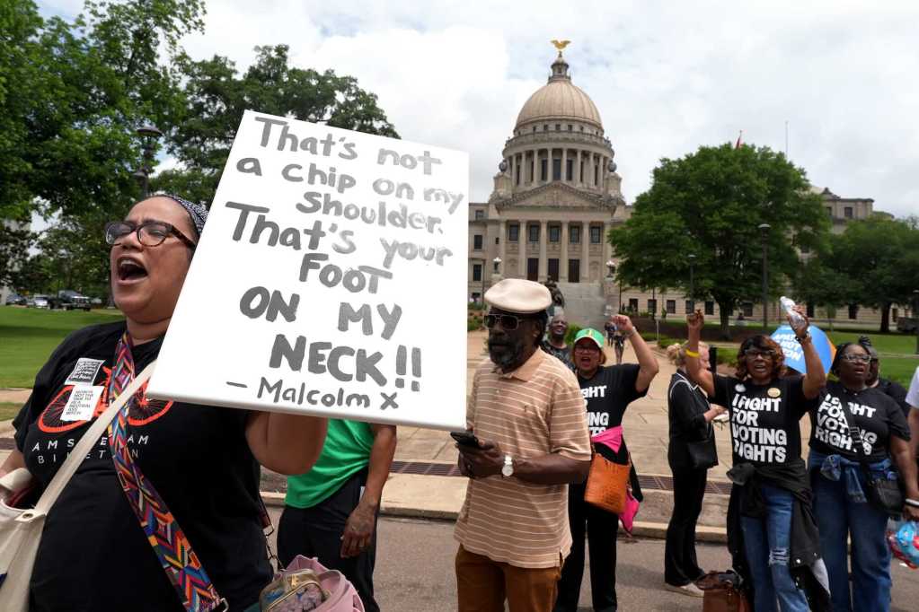 People march past the Mississippi capital building, some in shirts that read 'I Fight For Voting Rights'. A person holds a sign that reads 'That's not a chip on my shoulder, that's your foot on my neck!! - Malcom X'