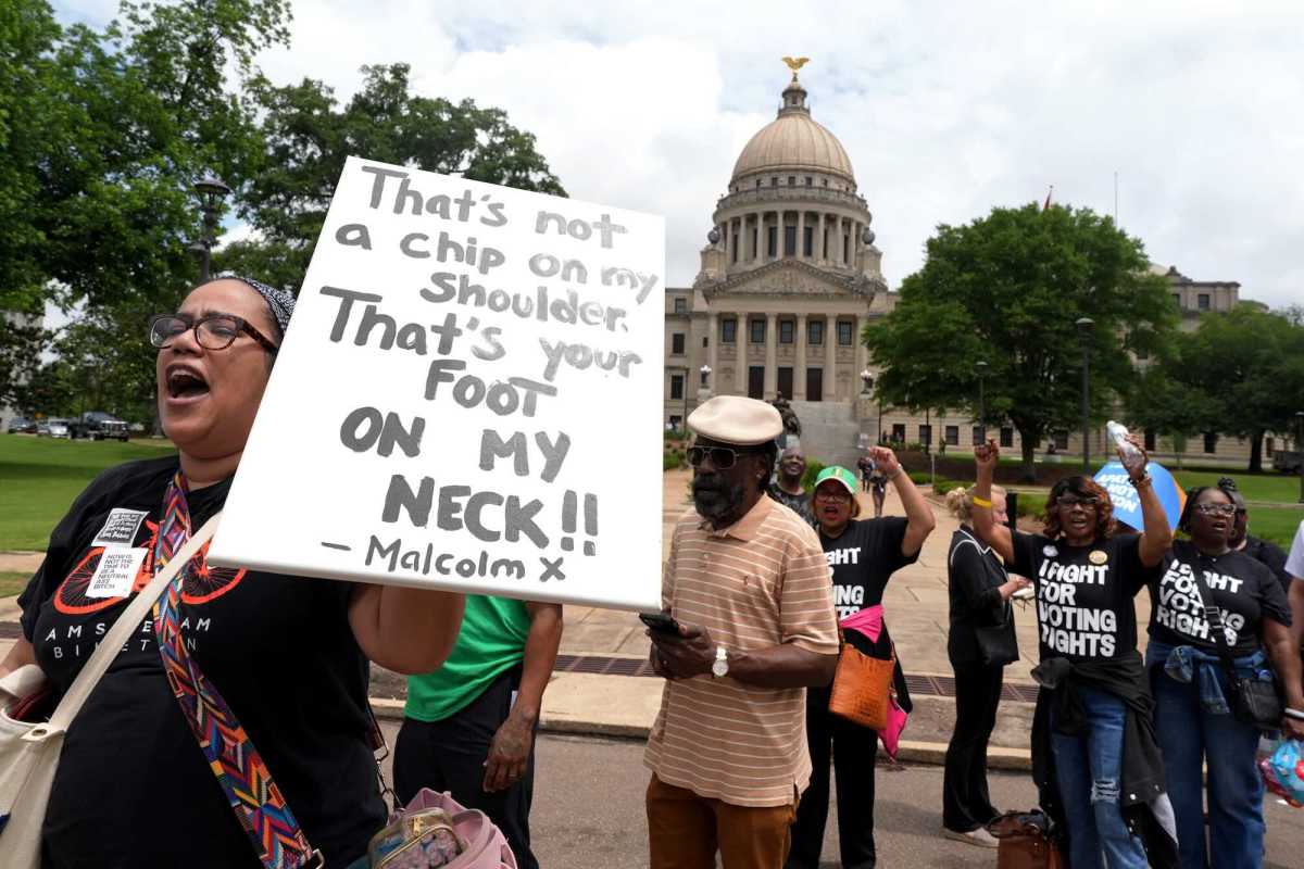 People march past the Mississippi capital building, some in shirts that read 'I Fight For Voting Rights'. A person holds a sign that reads 'That's not a chip on my shoulder, that's your foot on my neck!! - Malcom X'