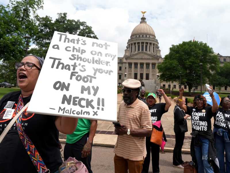 People march past the Mississippi capital building, some in shirts that read 'I Fight For Voting Rights'. A person holds a sign that reads 'That's not a chip on my shoulder, that's your foot on my neck!! - Malcom X'
