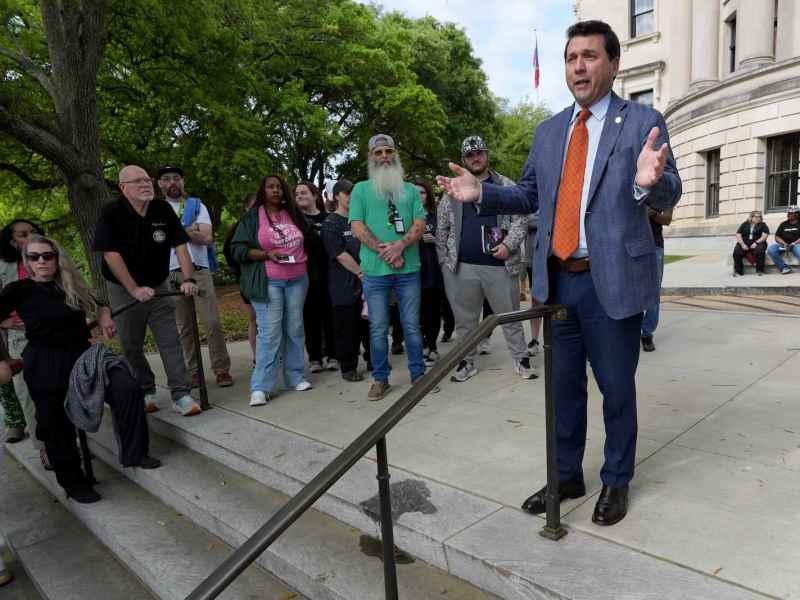 A man in a blue suit and orange tie speaks on the top of a set of outside stairs to onlookers