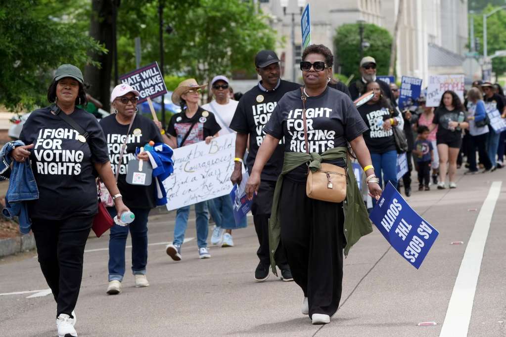 A line of people march down the road. Many are wearing black tshirts that read 'I Fight For Voting Rights'. Some hold signs like "Hate Will Not Make Us Great" and "As Goes The South'