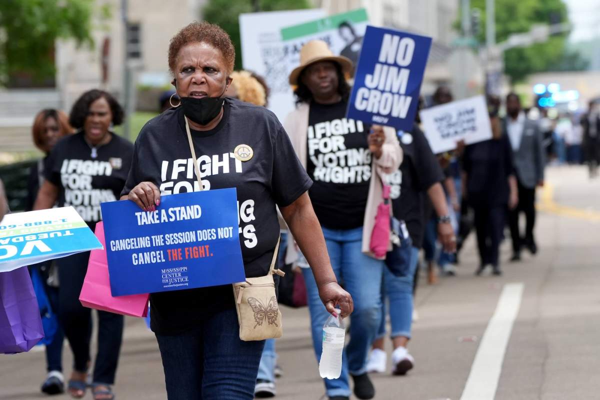 A line of people march down the road. Many are wearing black tshirts that read 'I Fight For Voting Rights'. Some hold signs like "No Jim Crow" and "Take a Stand. Cancelling the Session Does Not Cancel the Fight"