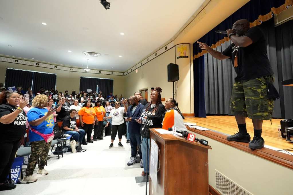 A man stands up on an auditorium stage and speaks to a crowd of listeners