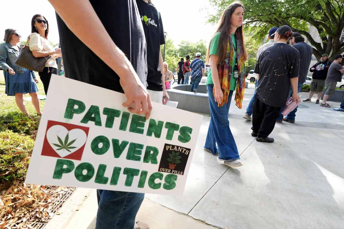 People gather outside, one holds a sign with "Patients Over Politics' written in green letters