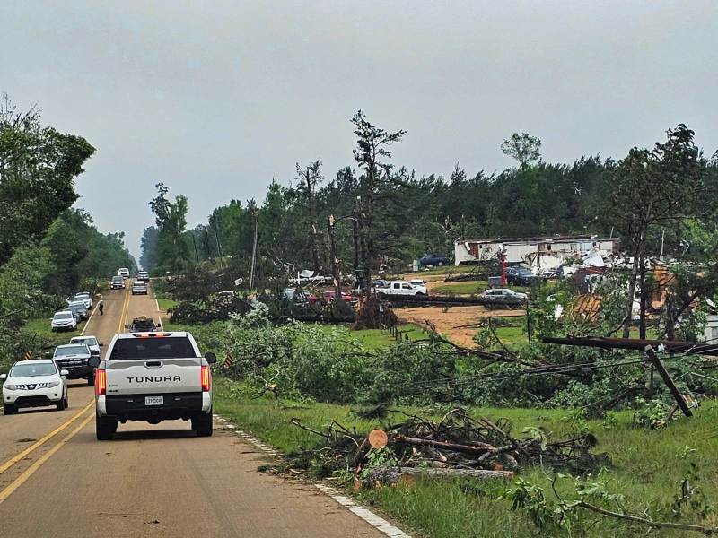 Traffic drives past buildings with tornado-damaged trees strewn about