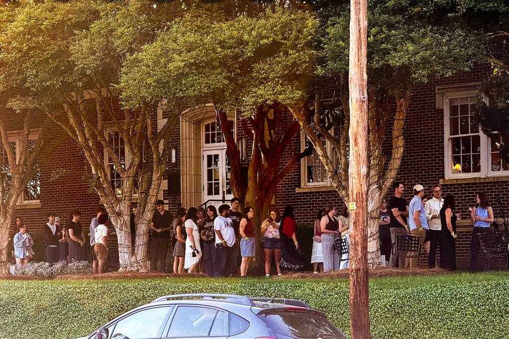 People stand in a long line under a row of trees outside of a brick building