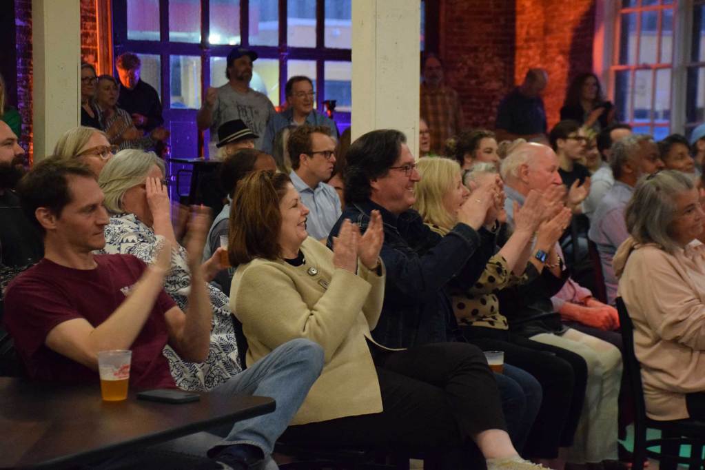 Seated audience members at an evening show in a brick building clap for the performers.