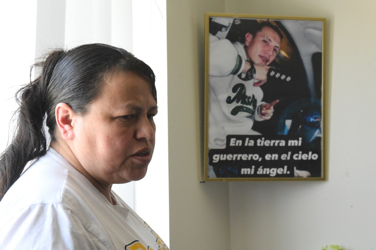 Closeup of a woman inside her home. A framed photo of her adult son hangs behind her with the words 'En la tierra mi guerrero, en el cielo mi ángel.'