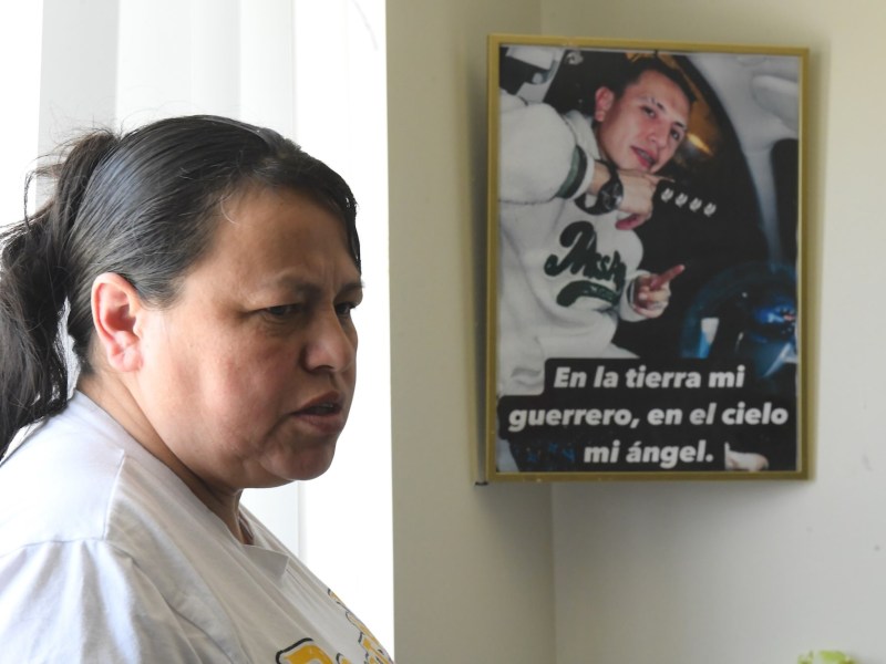 Closeup of a woman inside her home. A framed photo of her adult son hangs behind her with the words 'En la tierra mi guerrero, en el cielo mi ángel.'