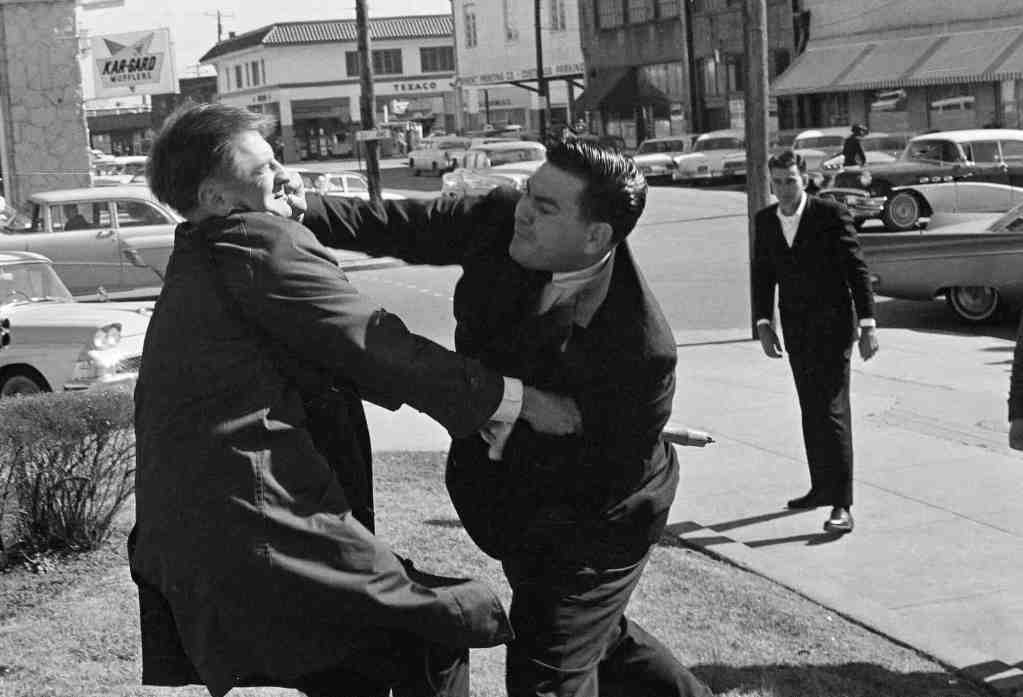 An old black and white photo from the 1960's showing two men in suits on a downtown sidewalk, throwing punches