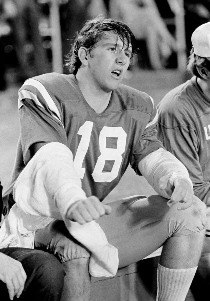 An old black and white photo from the 1970's of Archie Manning in football uniform, sitting on the sideline and grimacing as his arm is wrapped in a towel