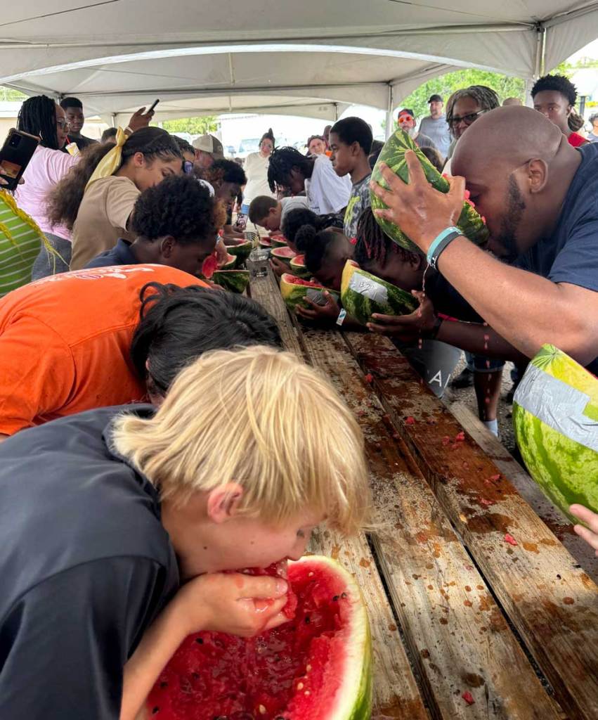 People lined up on both sides of a long wooden table, quickly eating watermelon in a contest