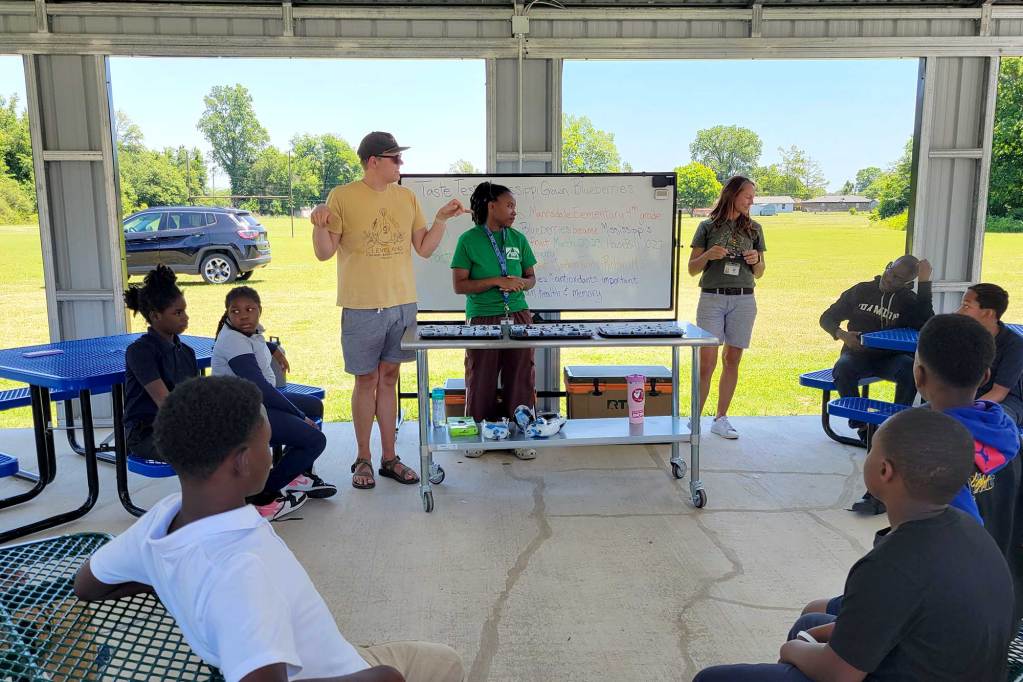 Three people stand at a desk and white board under an outside building, teaching children that sit on park benches