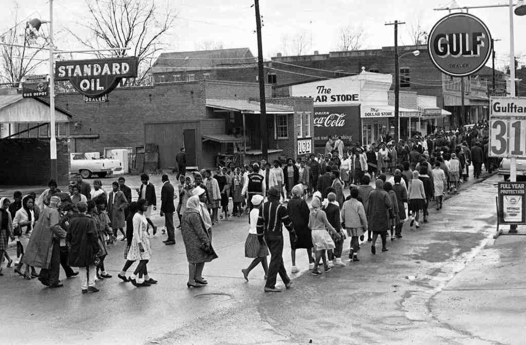 An old black and white photo from the 1960's showing hundreds of Black people marching up and down a main downtown road between "Standard Oil" and "Gulf"
