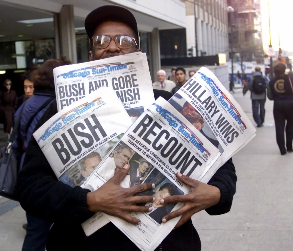 A man stands on a busy sidewalk with arms full of Chicago Sun-Times newspapers with four different large headlines 'Bush and Gore' 'Bush' "Recount' 'Hillary Wins'
