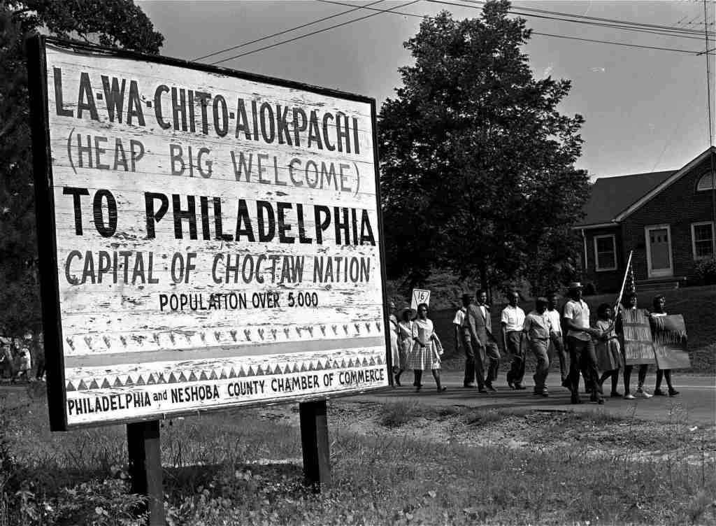 An old black and white photo from the 1960's of Black people marching down the road. In the foreground is a large white sign that reads 'LA-WA-CHIFO-ALOKPACAI (HEAP BIG WELCOME) TO PHILADELPHIA CAPITAL OF CHOCTAW NATION POPULATION OVER 5.000 PHILADELPHIA and NESHOBA COUNTY CHAMBER OF COMMERCE' POPULATTON OVER 5.000 PHILADELPHIA and NESHOBA COUNTY CHAMBER OF COMMERCE'
