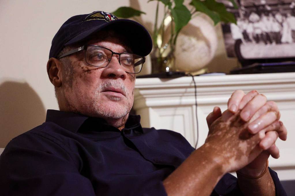 A closeup of Cliff Baptiste, wearing a baseball cap and button down shirt, seated in his living room
