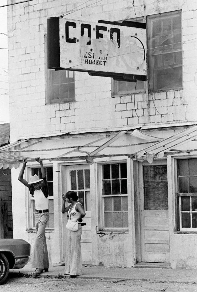 An old black and white photo from the 1970's of two people standing outside of a damaged white building