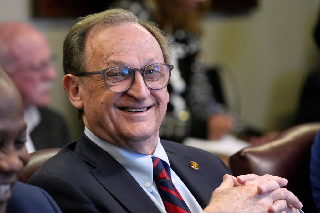 A man in a suit with blue and red striped tie leans back, smiling during a meeting
