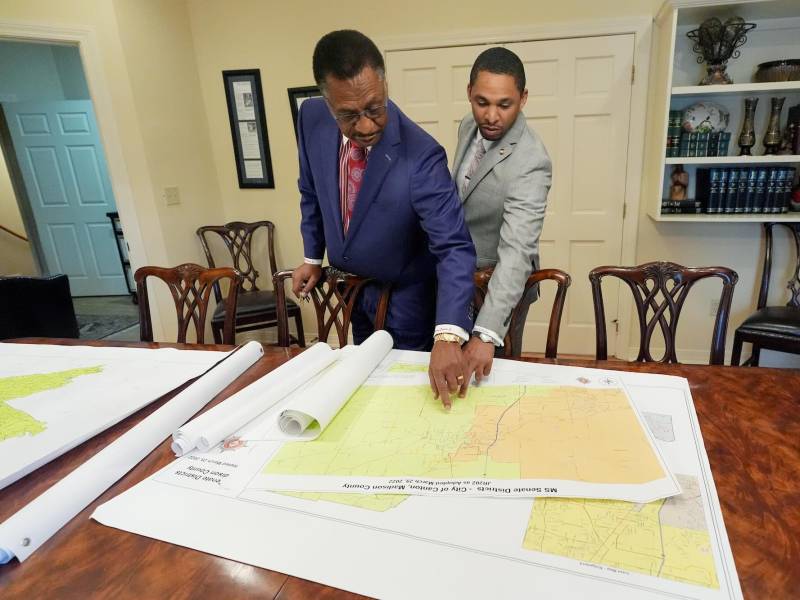 Edward and Bradford Blackmon stand at a table in an office, pointing at something on the first of several stacked maps