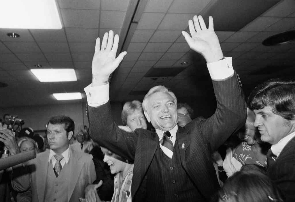 An old black and white photo from the 1970's showing a man in a dark suit with both hands up in celebration among a crowd of people
