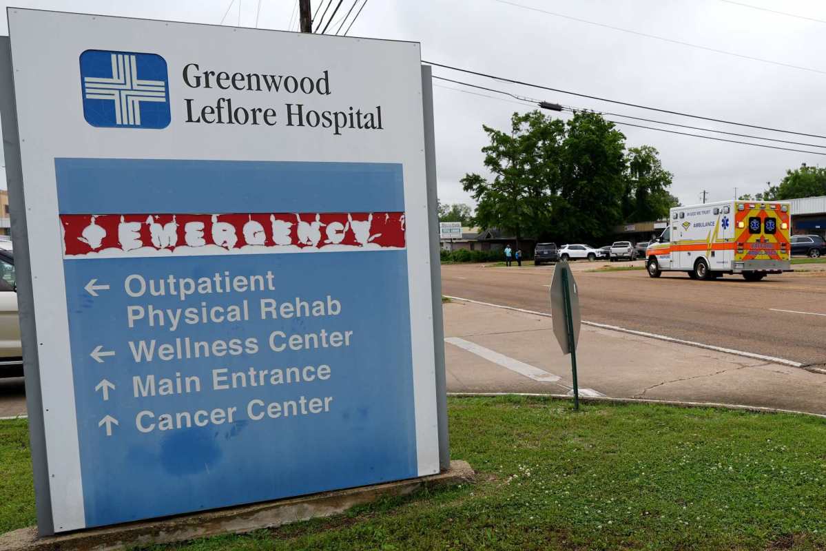 A faded blue and white Greenwood Leflore Hospital sign by the road. An ambulance can be seen driving down the road beyond.