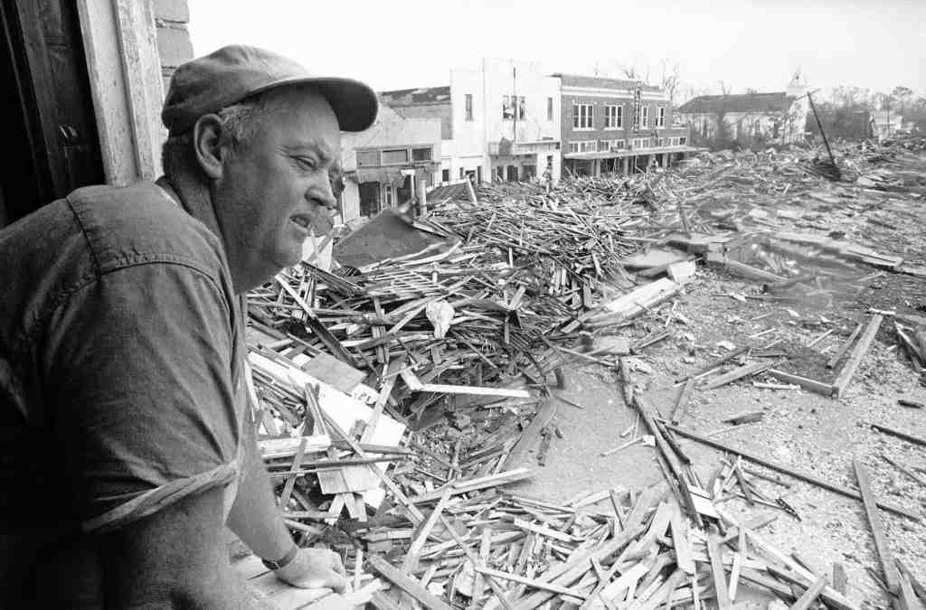 An old black and white photo from the 1960's of a man looking down from a building window at the massive debris left in the street from a hurricane