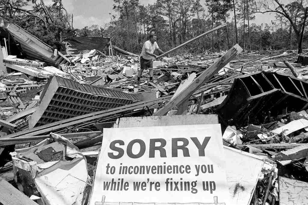 An old black and white photo from the 1960's showing a man in the center of a huge pile of hurricane destroyed building debris, tossing a wooden board to the side