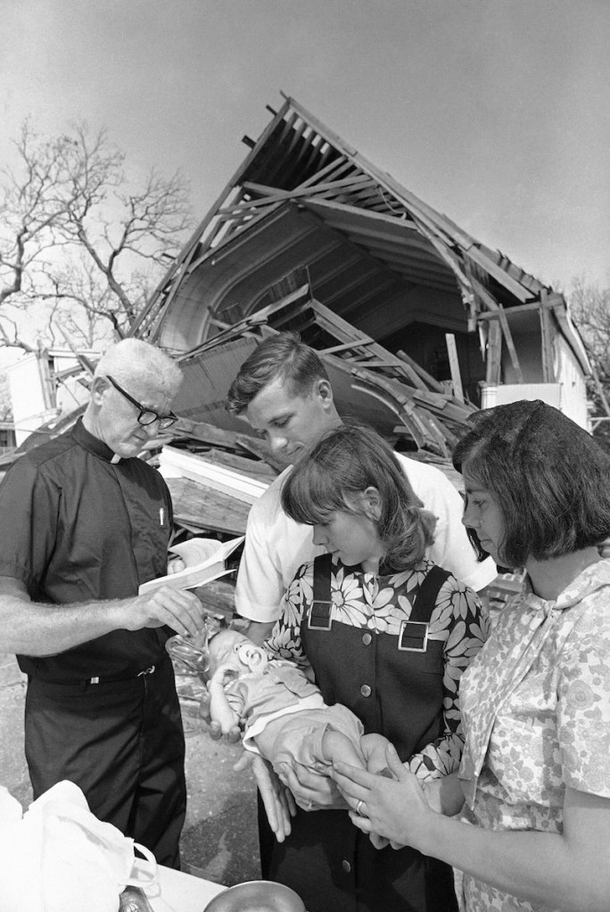 An old black and white photo from the 1960's showing a family getting their infant baptized outside in front of a hurricane-destroyed church