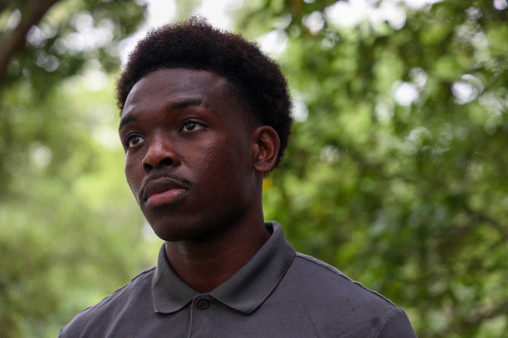 A closeup of Israel Makoka in a grey collared shirt, standing outside with greenery all behind him