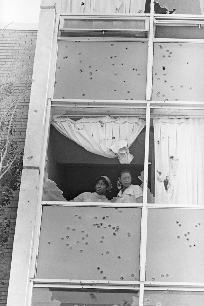 An old black and white photo from the 1970's showing two women look through an upstairs dorm window, the whole side of the building riddled with bullet holes