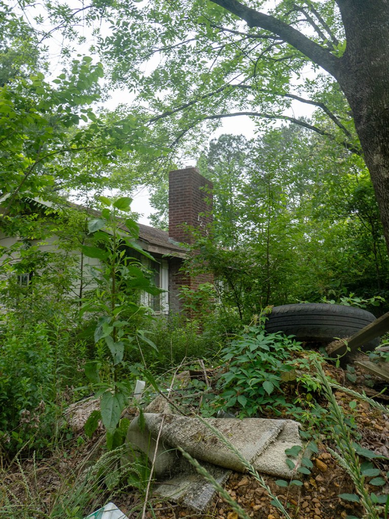 A home sits in an overgrown lot, trash and other debris piling up outside