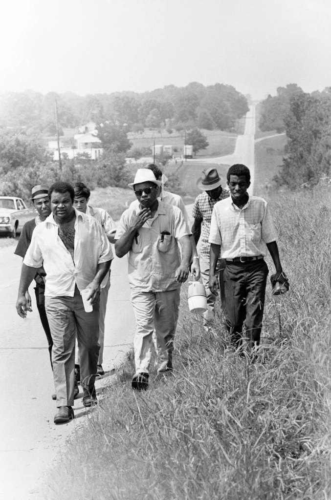 An old black and white photo from the 1960's of James Meredith and 6 other men walking down the side of a road
