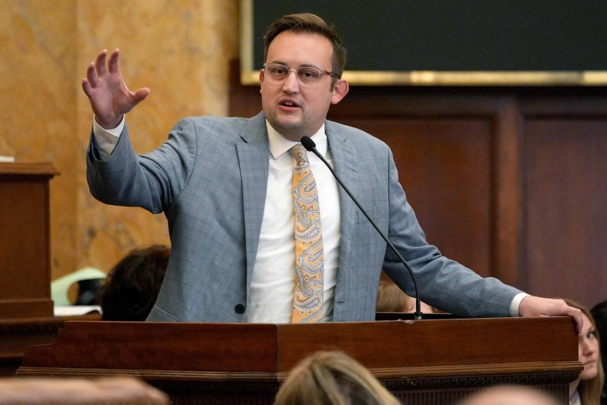 Jansen Owen, in a grey suit and yellow paisley tie, speaking at a mic during a capital meeting with one arm gesturing widely