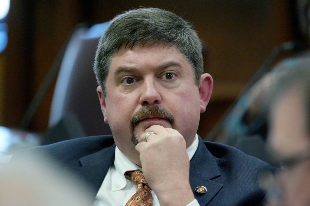A closeup of Jeremy England sitting in the Capitol, hand resting on his chin as he watches people talk