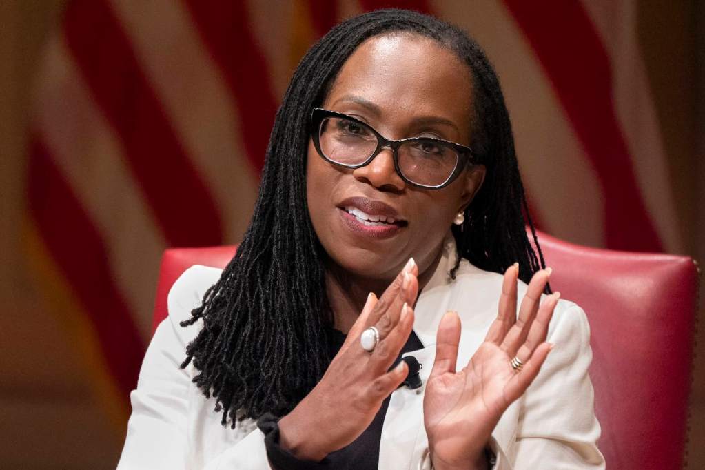 Kentaji Brown, seated at a meeting wearing a white jacket and speaking, gesturing with both hands