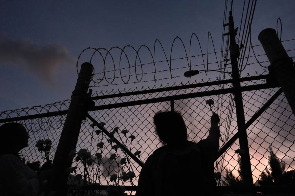 A woman holds a flower up toward a barbed-wire fence at night.