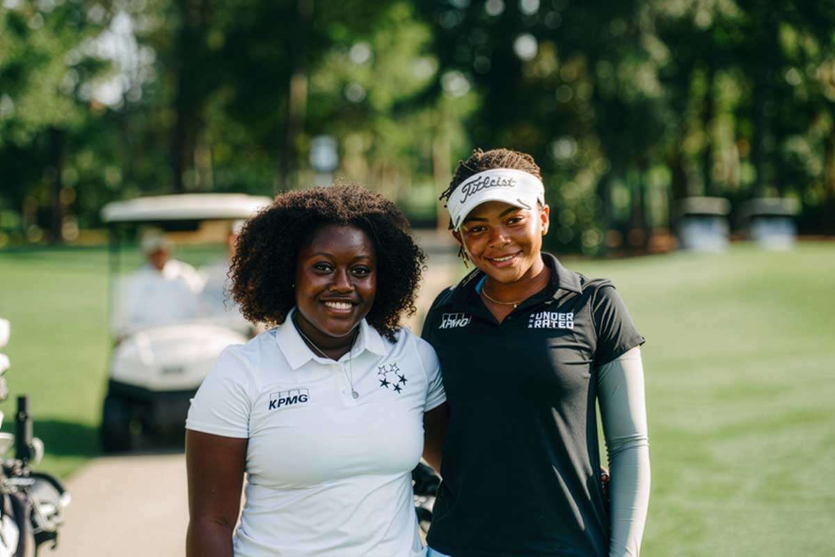 Two women wearing gold shirts that reads 'KPMG Underrated' stand together on a golf course