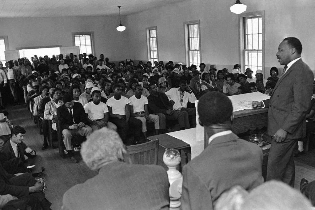 An old black and white photo from the 1960's showing Martin Luther King speaking to a room full of seated people