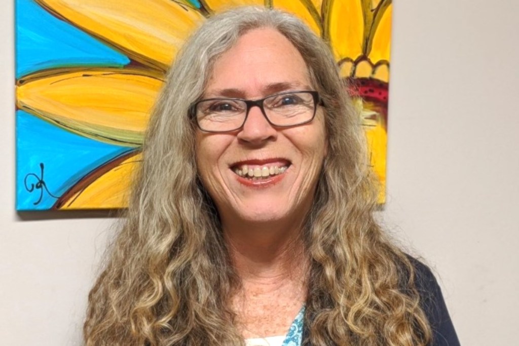 A woman with long wavy blonde hair and glasses smiles in front of a painting of a sunflower