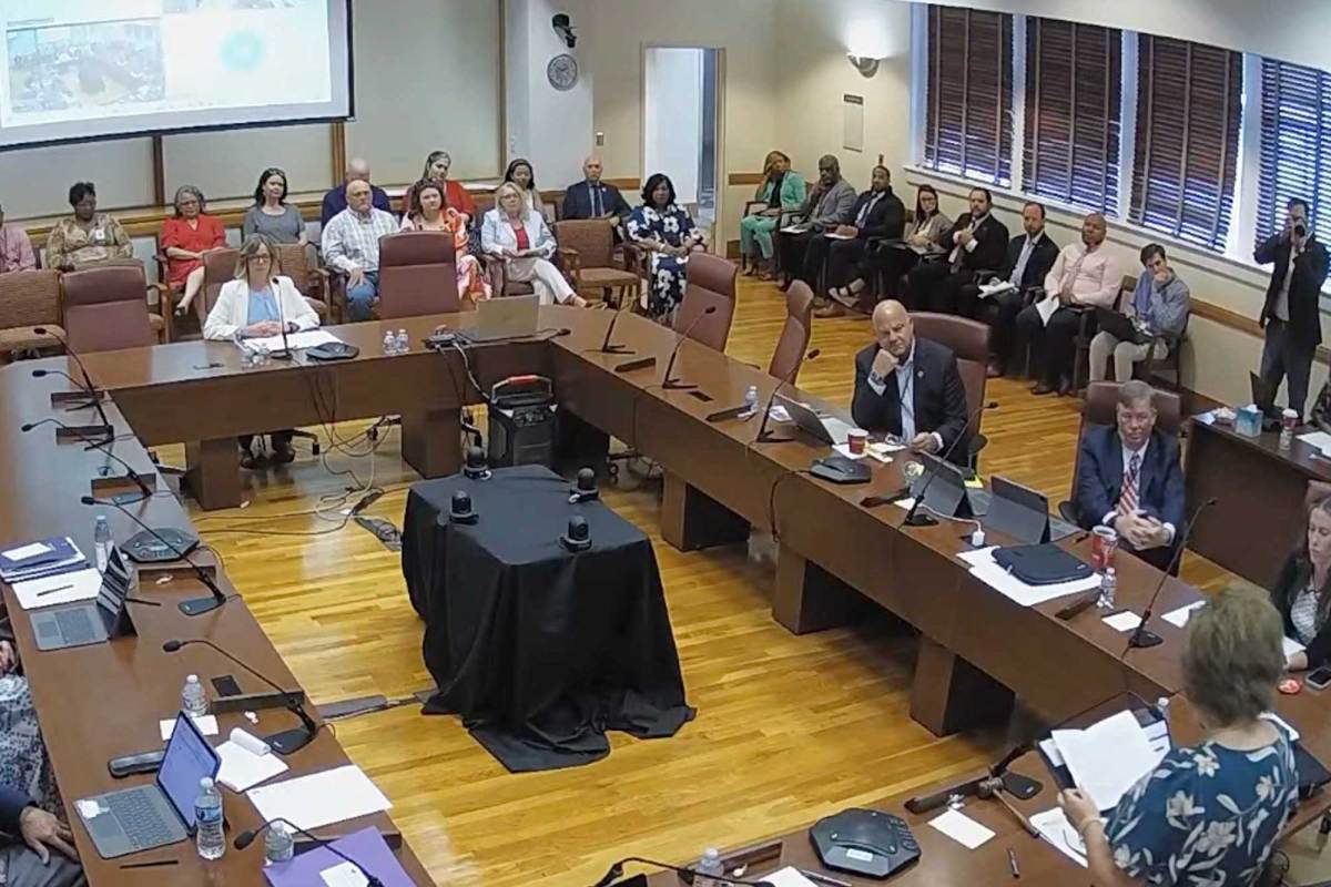 A room with people sitting at conference tables that form a square, and others listening from the edges of the room
