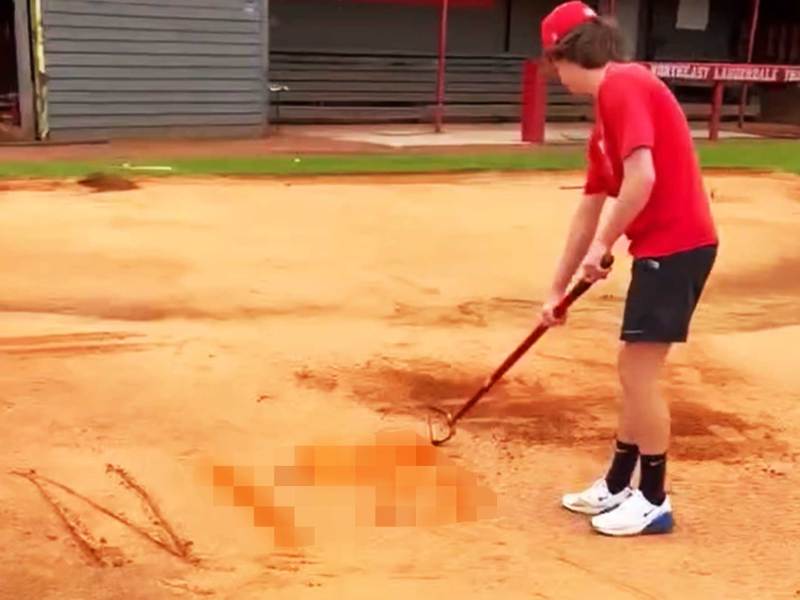 A teen in a red and black baseball outfit scratching a word in the dirt of the sports field