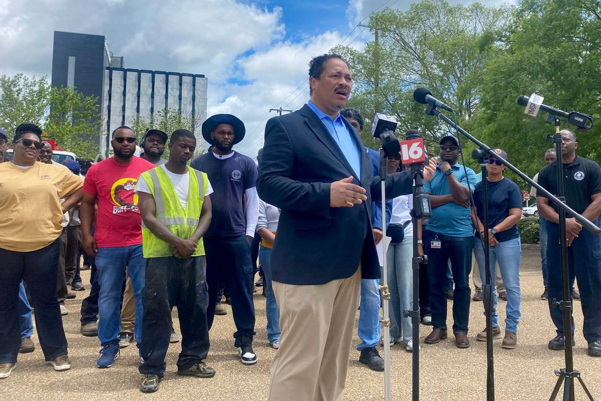 A man in a suit speaks at a microphone with other people standing behind him during an event held outside