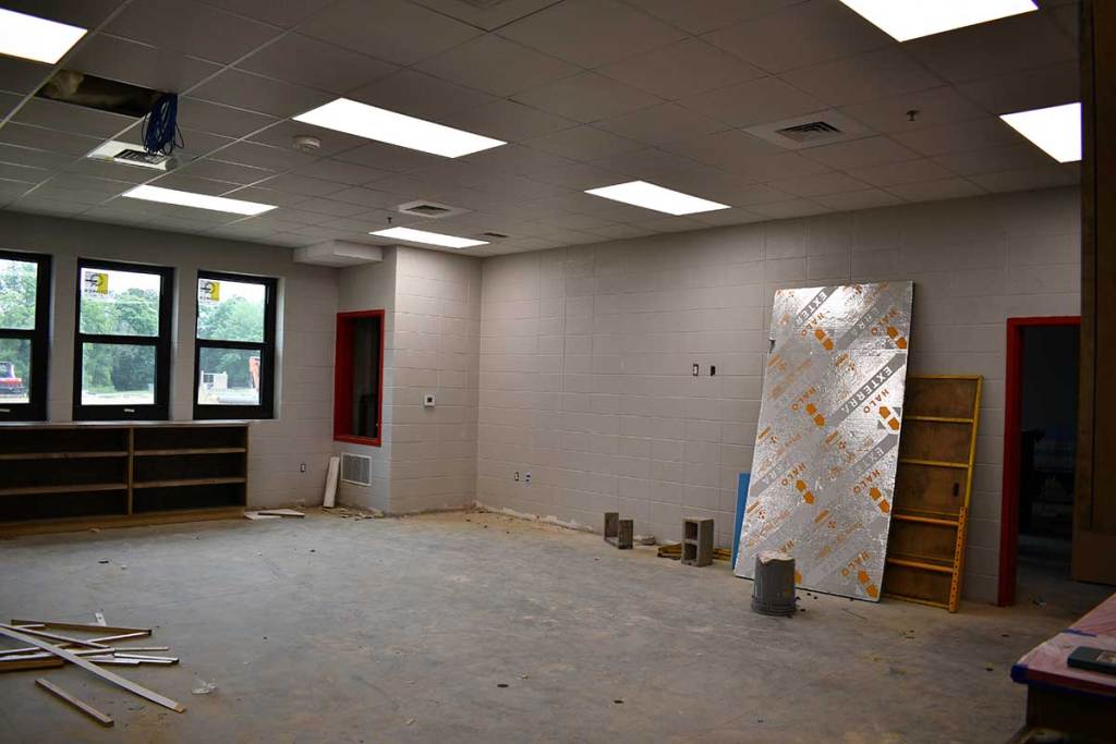 An unfinished classroom has bare floors, ceiling lights, windows and construction materials inside.