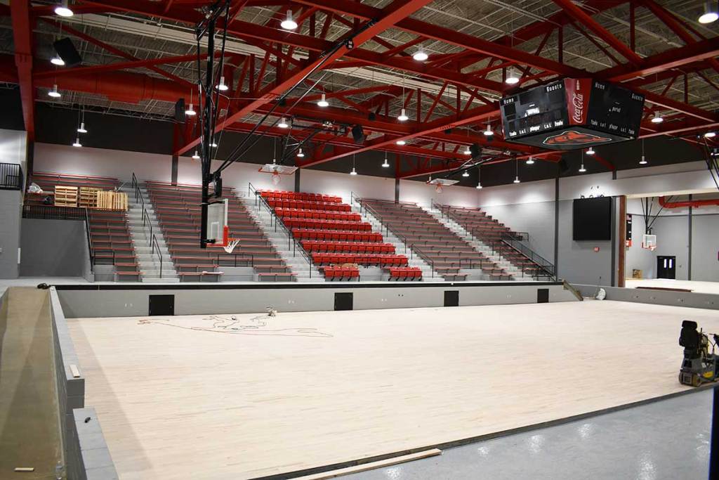 A large school gymnasium has red seating, basketball goals, a stage and construction equipment on the floor.