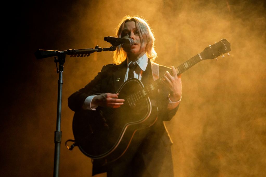 Musician Phoebe Bridgers stands on a dark stage lit with orange fog, strumming a guitar and singing into a microphone